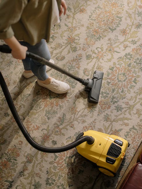 A professional cleaner dressed in a white protective suit, gloves, and blue shoe covers is using a vacuum cleaner with a long hose attachment to deep clean a beige carpet in a well-lit, modern living