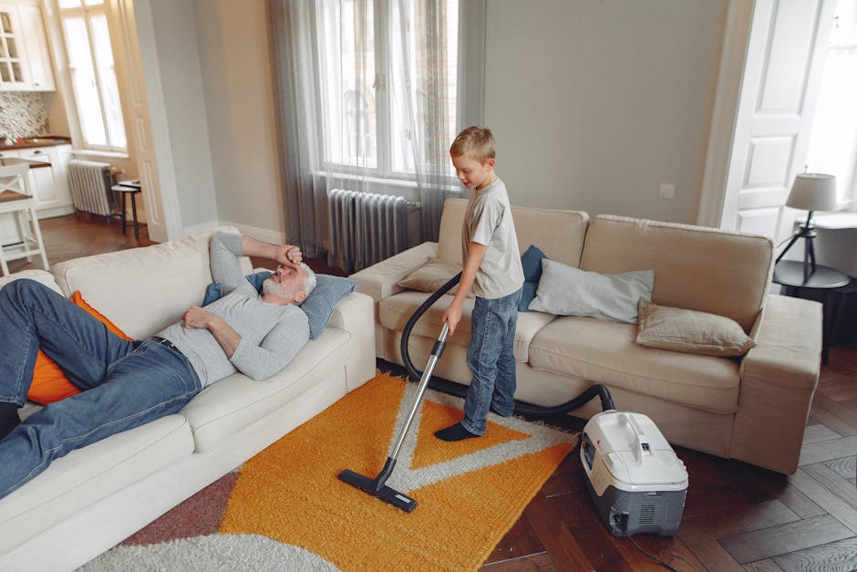 A young boy vacuuming a beige sofa in a living room with natural light coming through large windows, while an older man with gray hair and a beard reclines on the sofa, resting with his eyes closed an